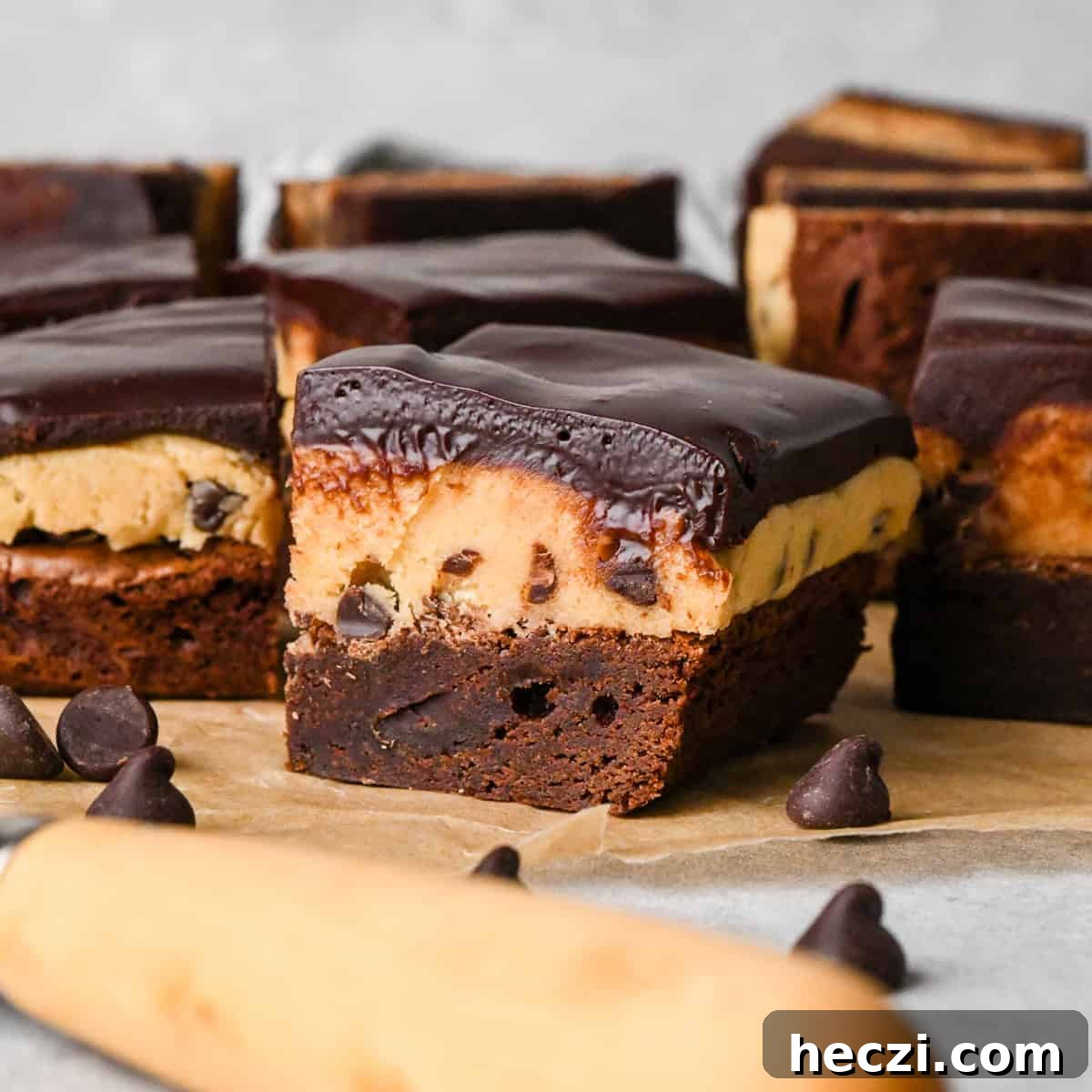 3 cookie dough brownies with a fudgy chocolate brownie on the bottom, edible chocolate chip cookie dough in the middle, and a glossy chocolate ganache on top on parchment paper. Surrounded by other cookie dough brownies and chocolate chips