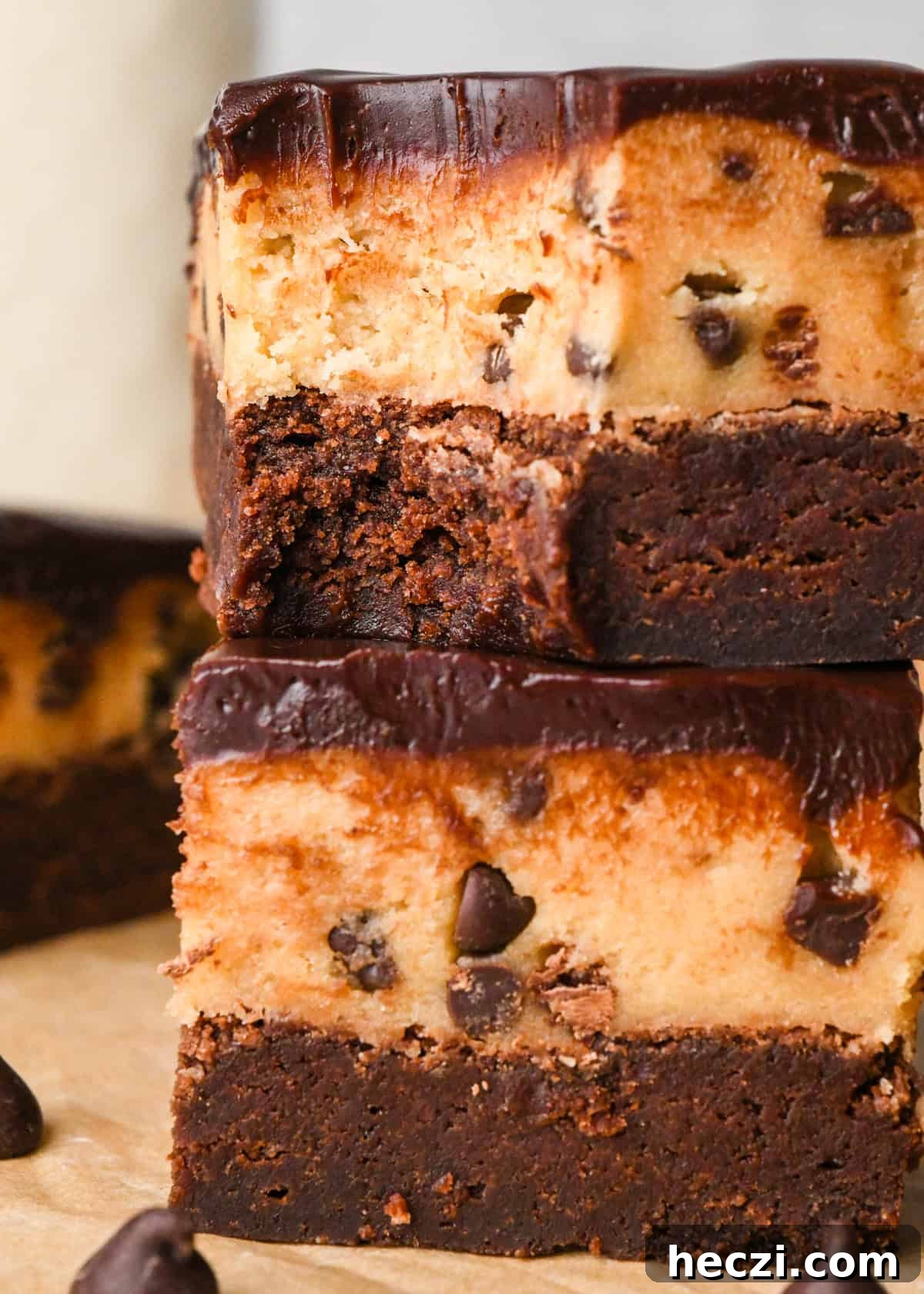 close up photo of a stack of two cookie dough brownies with a fudgy chocolate brownie on the bottom, edible chocolate chip cookie dough in the middle, and a glossy chocolate ganache on top on parchment paper. The top brownie has a bite taken out fo it.  Surrounded by other cookie dough brownies and chocolate chips