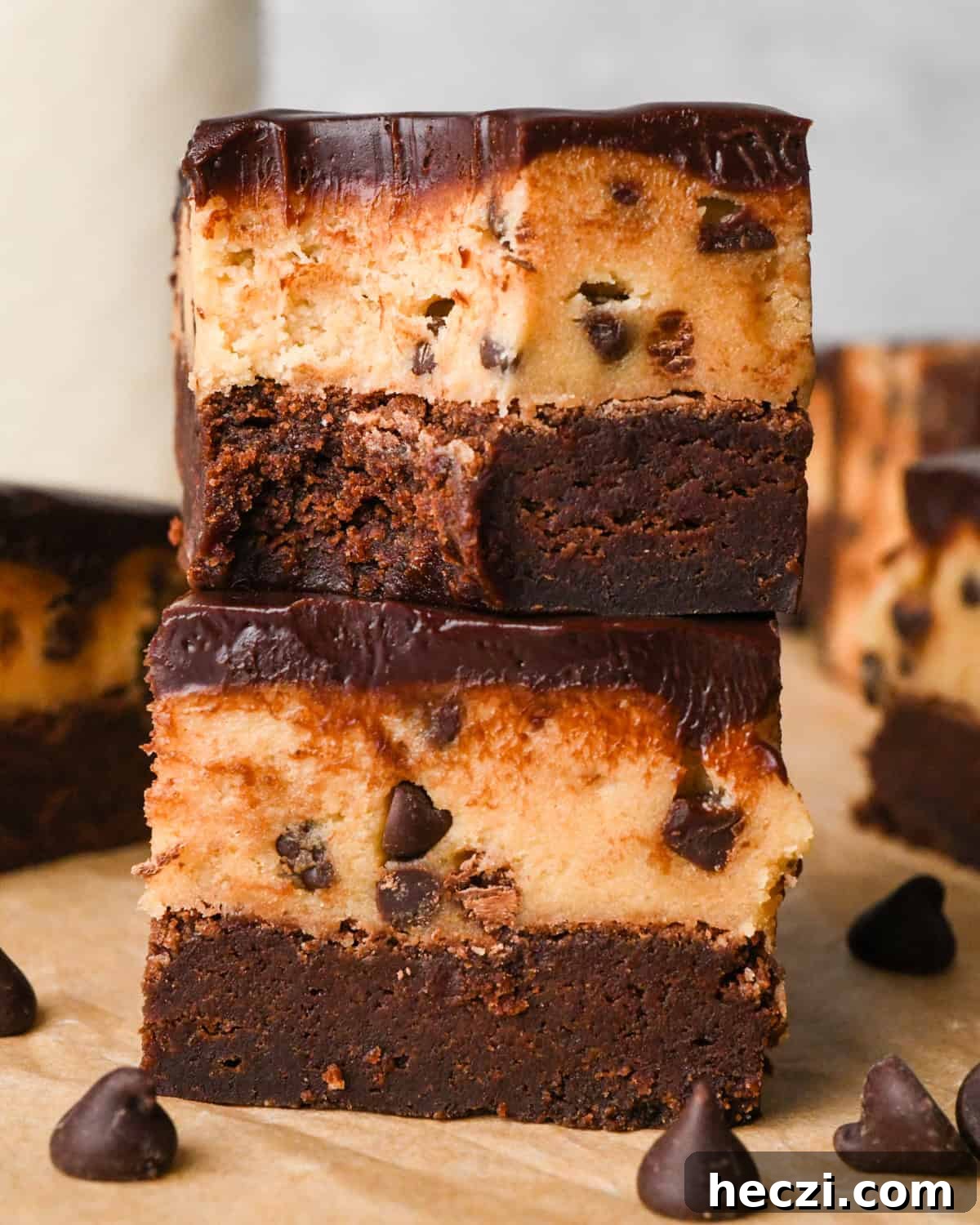 stack of two cookie dough brownies with a fudgy chocolate brownie on the bottom, edible chocolate chip cookie dough in the middle, and a glossy chocolate ganache on top on parchment paper. The top brownie has a bite taken out fo it.  Surrounded by other cookie dough brownies and chocolate chips