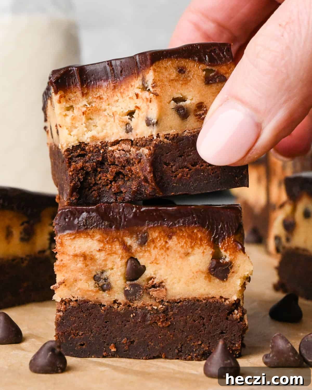 a stack of two cookie dough brownies with a fudgy chocolate brownie on the bottom, edible chocolate chip cookie dough in the middle, and a glossy chocolate ganache on top on parchment paper A hand is picking up the top brownie which has a bite taken out of it. Surrounded by other cookie dough brownies and chocolate chips