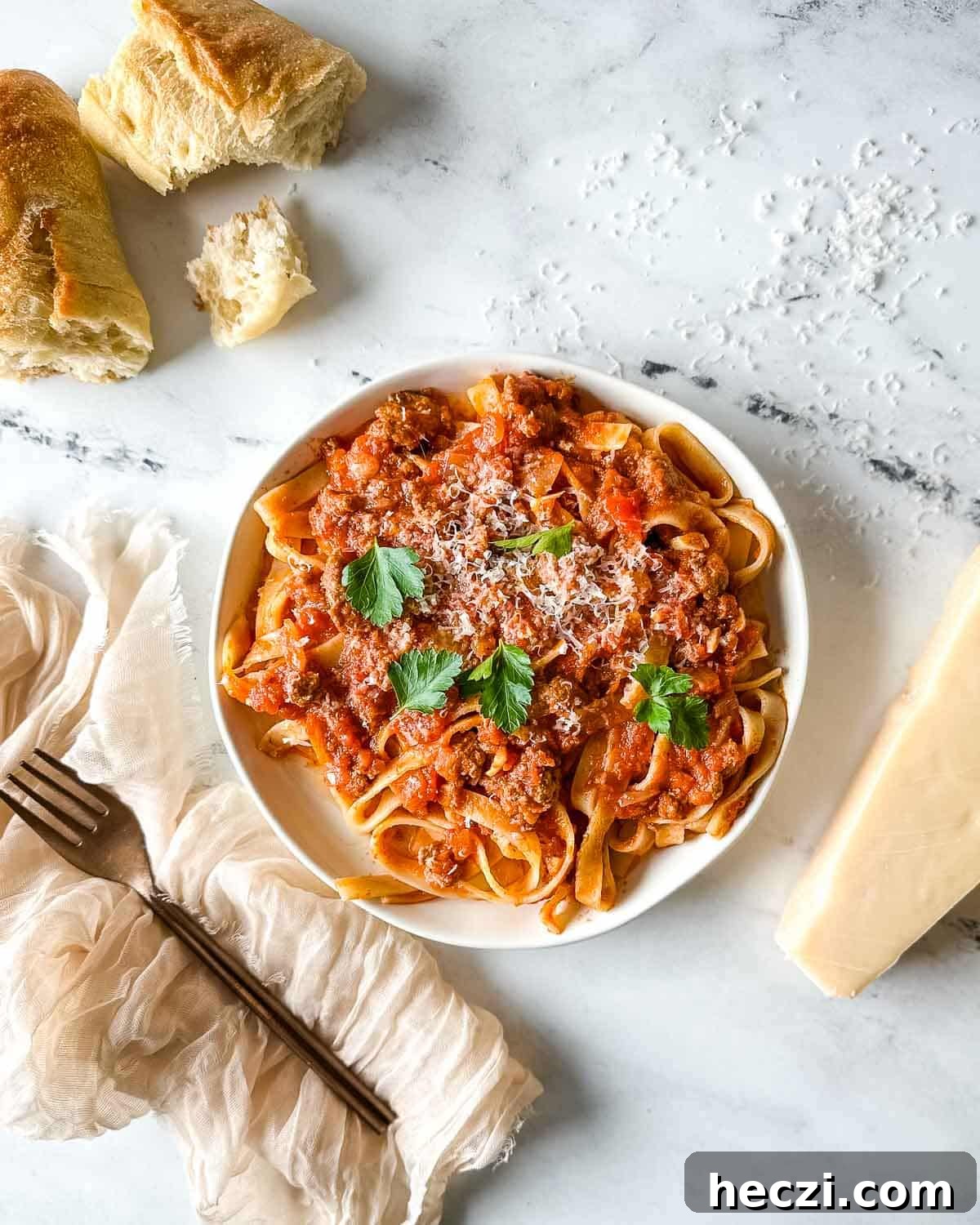 Slow Cooker Lamb Ragu: Tender Rustic Pasta Sauce Recipe 2 a plate of slow cooker lamb ragu over pappardelle pasta surrounded by a block of parmesan, a white linen, and torn baguette.