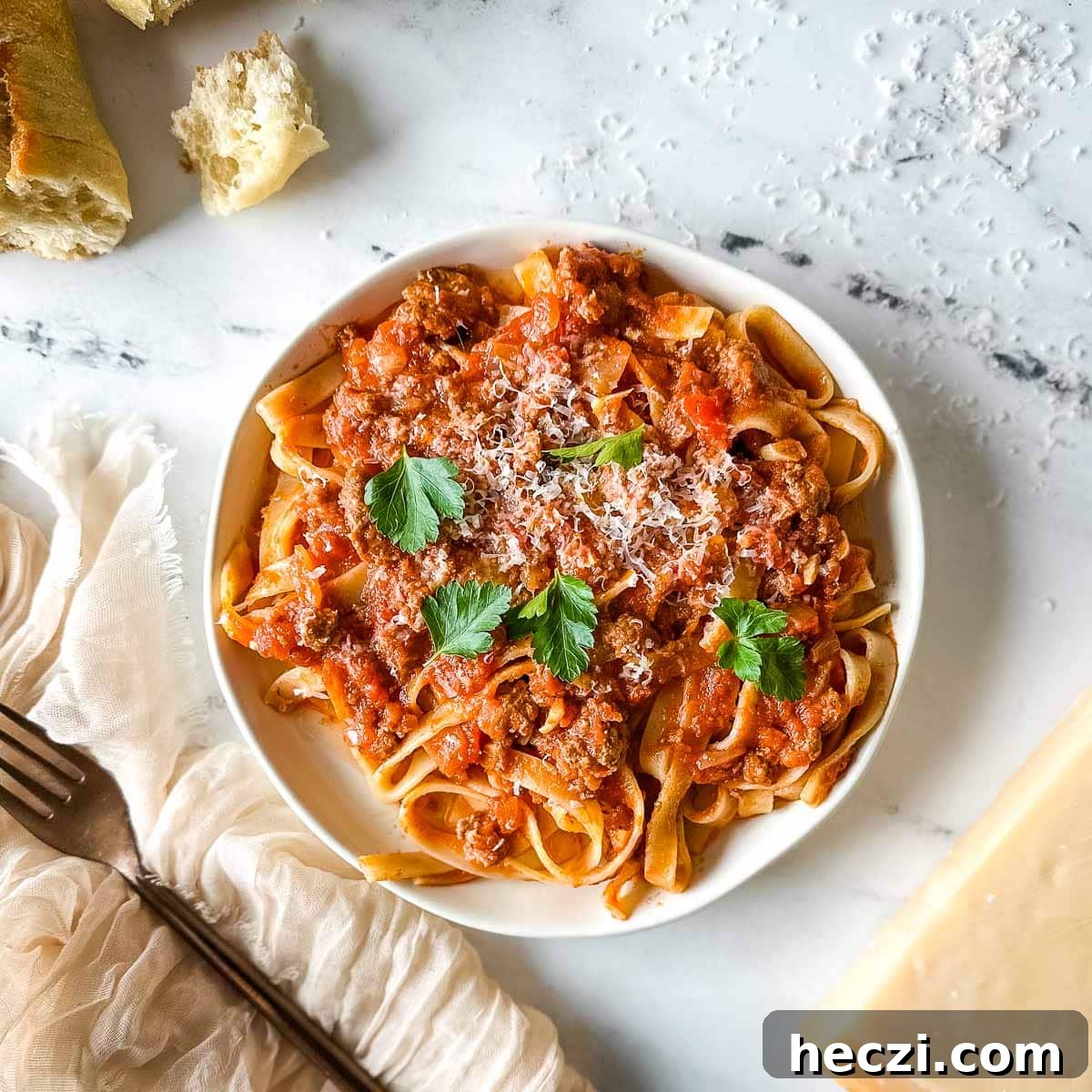 Slow Cooker Lamb Ragu: Tender Rustic Pasta Sauce Recipe 15 a plate of slow cooker lamb ragu over pappardelle pasta surrounded by a block of parmesan, a white linen, and torn baguette.