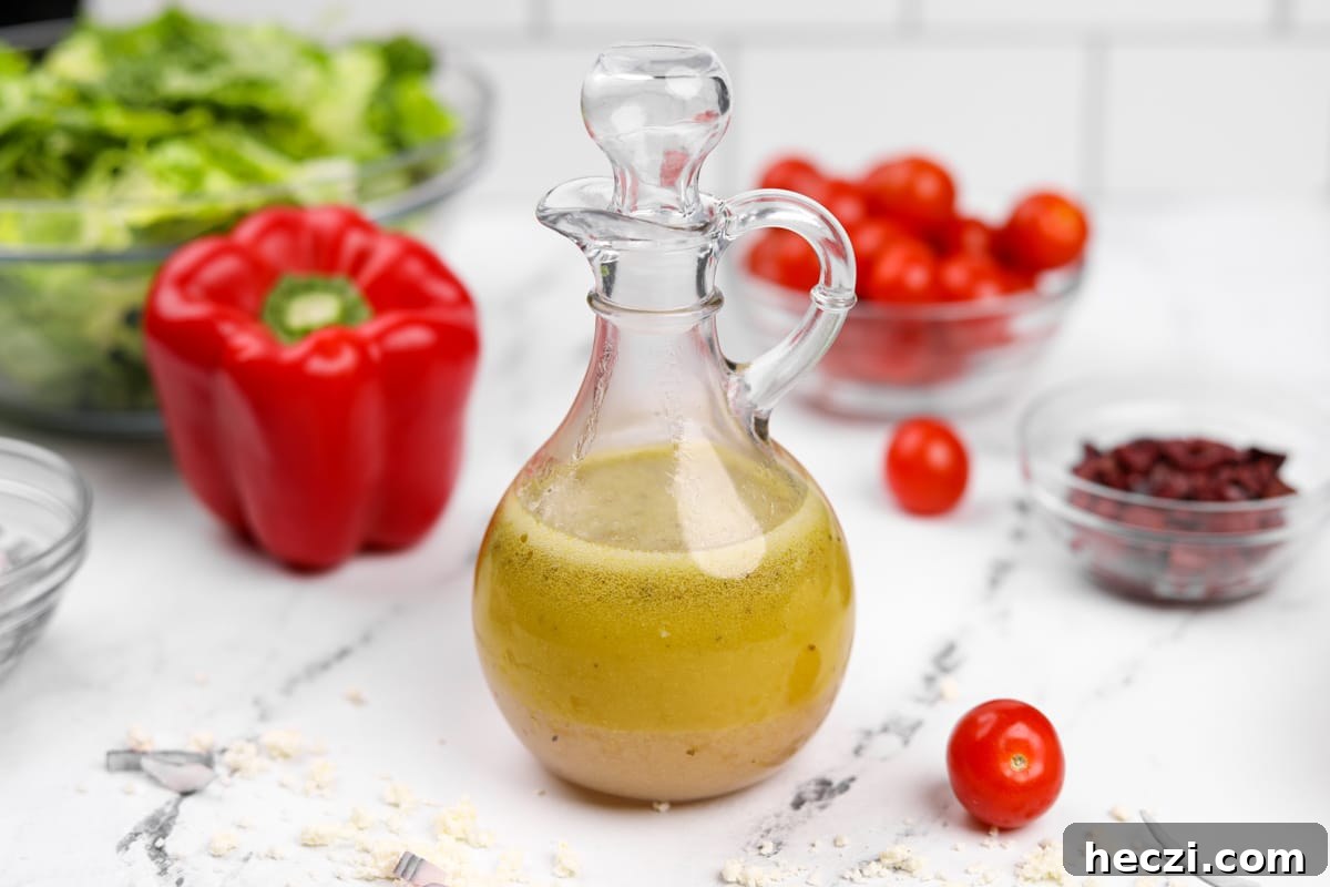 Authentic Greek Vinaigrette Recipe for Salads and Marinades 7 A glass container holding homemade salad dressing, with red pepper, lettuce, and tomato in the background.
