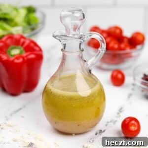 Authentic Greek Vinaigrette Recipe for Salads and Marinades 8 A glass cruet filled with homemade salad dressing, with red pepper, tomatoes, and lettuce in the background.