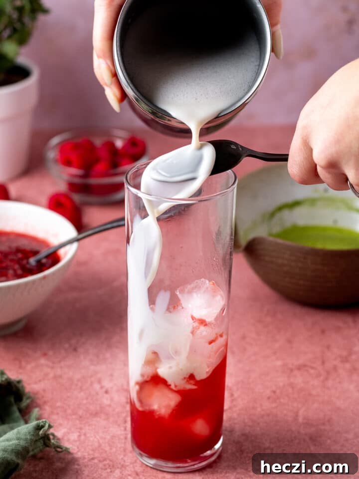 pouring frothed oat milk over a spoon into the glass with raspberry puree