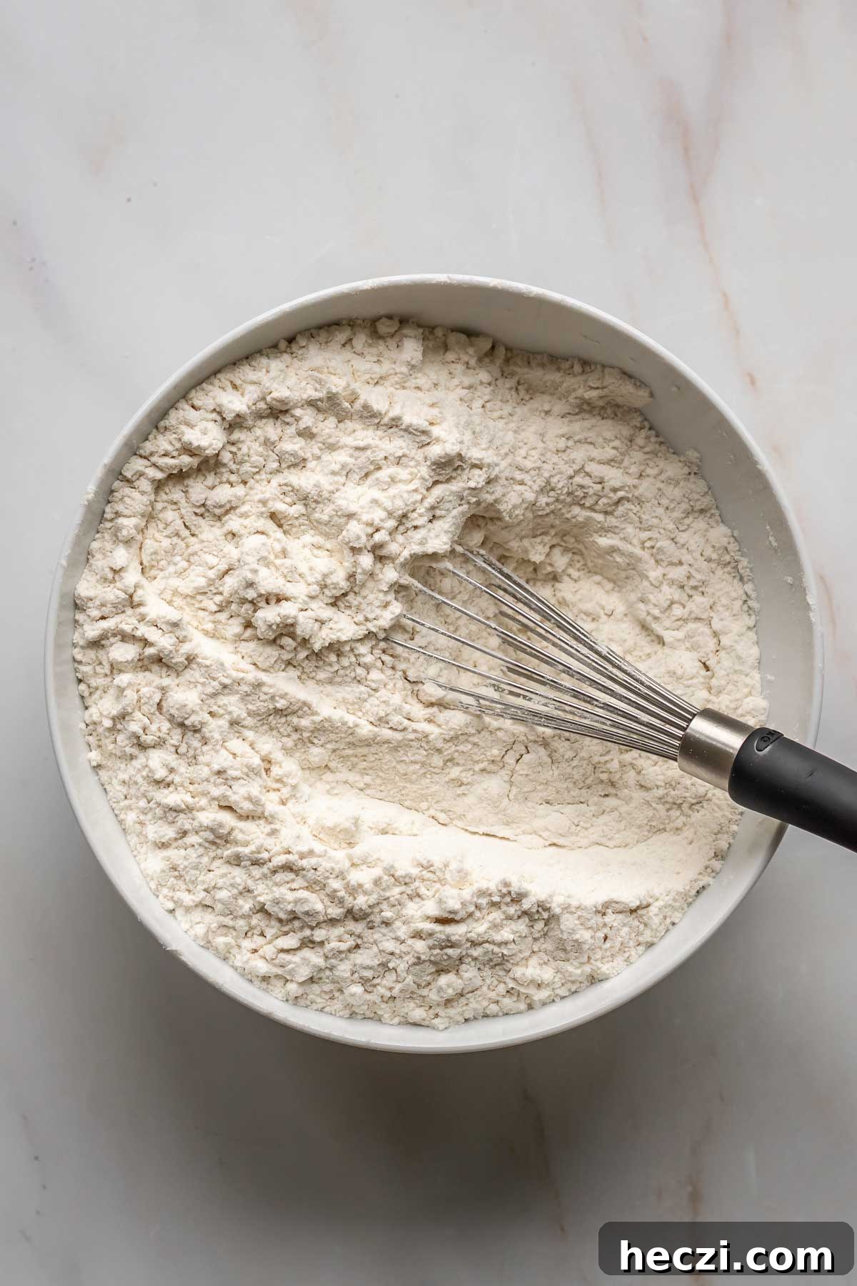 A whisk sits in a bowl of flour and dry ingredients.
