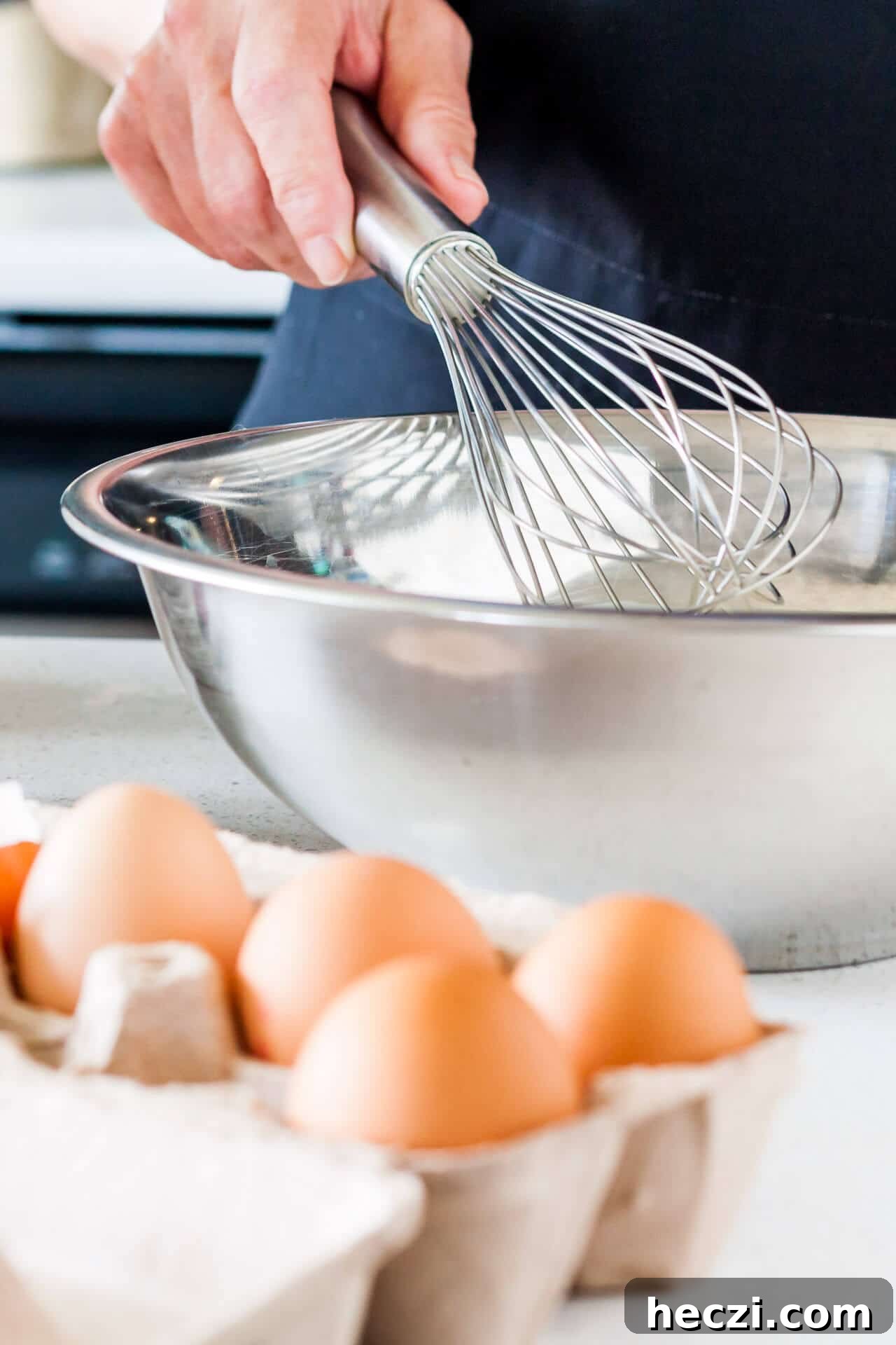 Whisking eggs by hand with a stainless steel whisk in a stainless steel bowl with a carton of brown eggs displayed next to the bowl