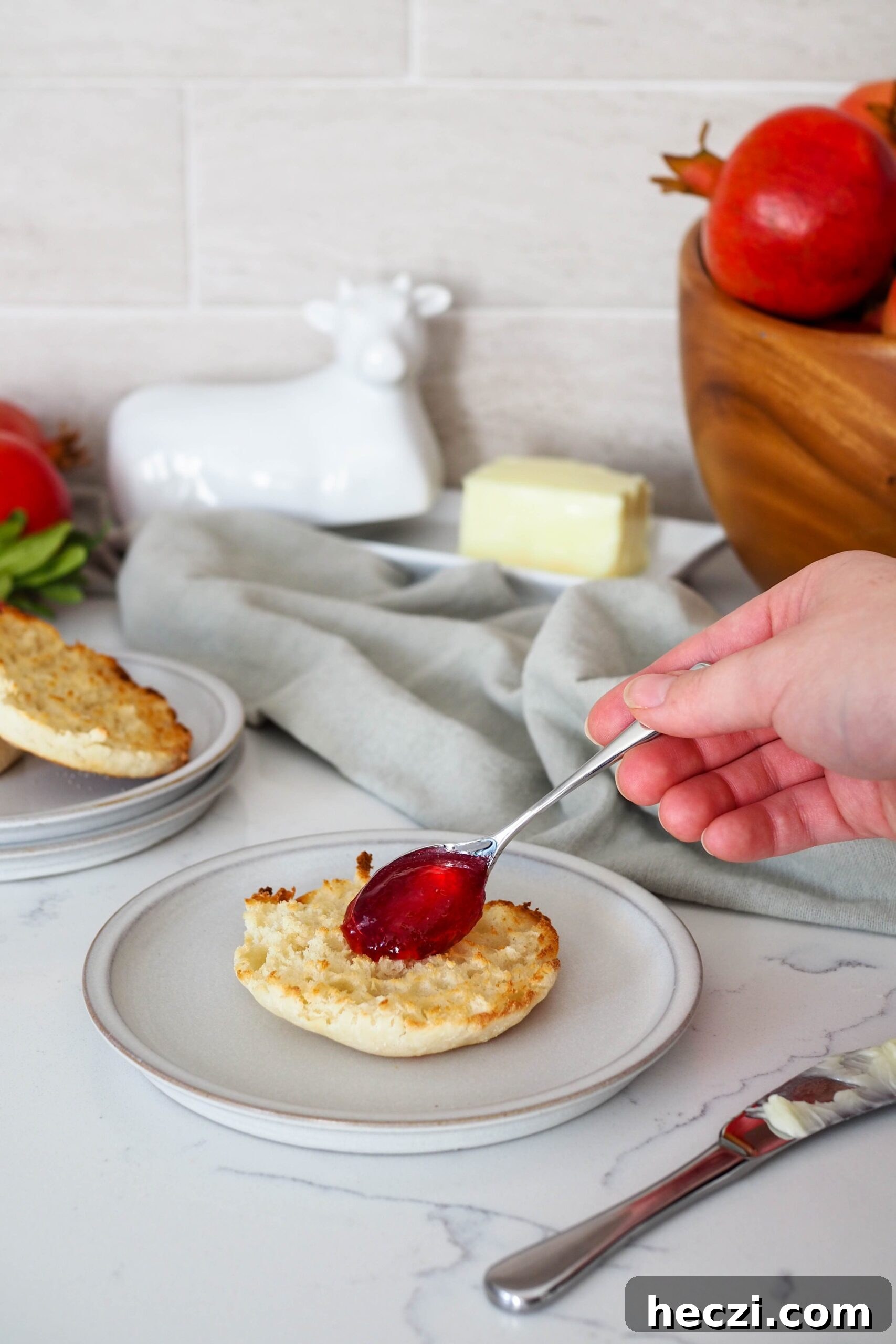 A small spoon spreads pomegranate jelly onto an English muffin.