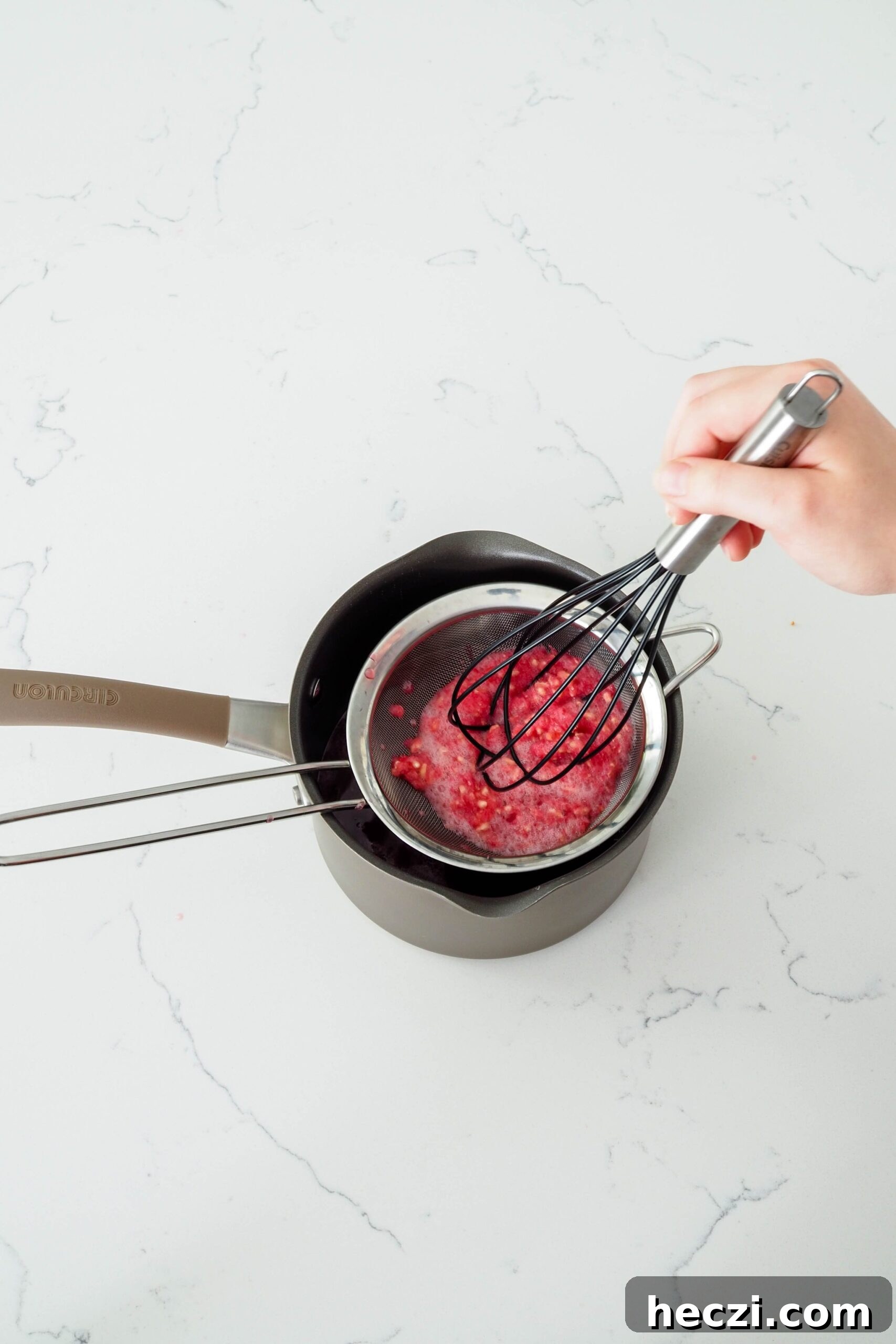 A hand pushes a whisk into the pomegranate arils in a fine mesh strainer.
