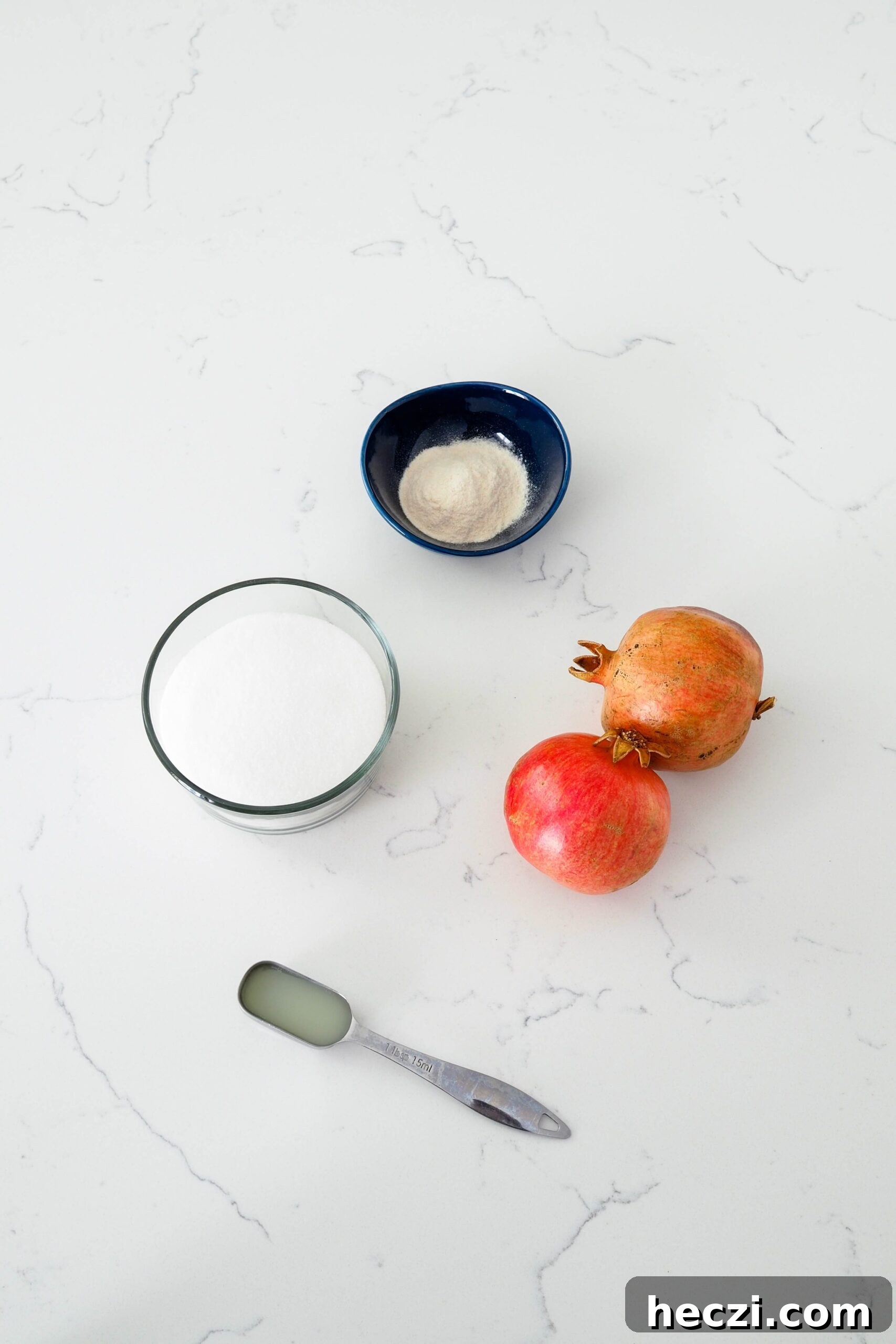 Two pomegranates, lemon juice, sugar, and pectin on a white counter.