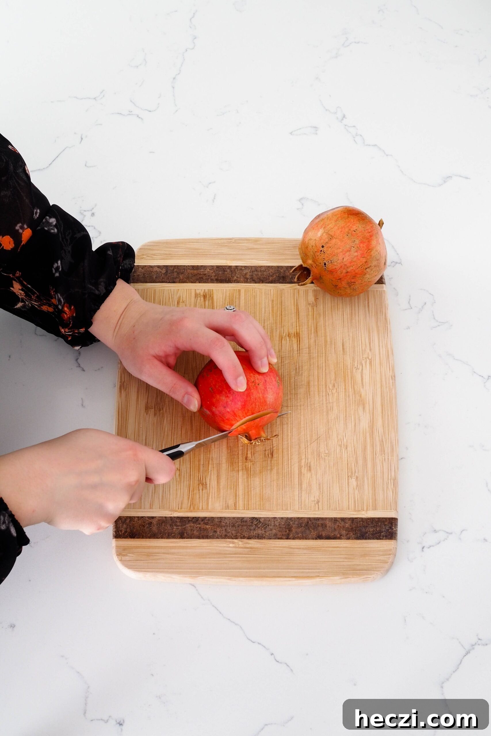 The crown of a pomegranate is being sliced off on a cutting board with a paring knife.