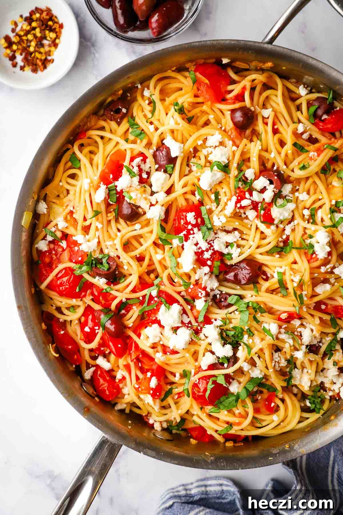 Overhead view of a pan of spagetti with tomatoes, olives and feta garnished with basil.