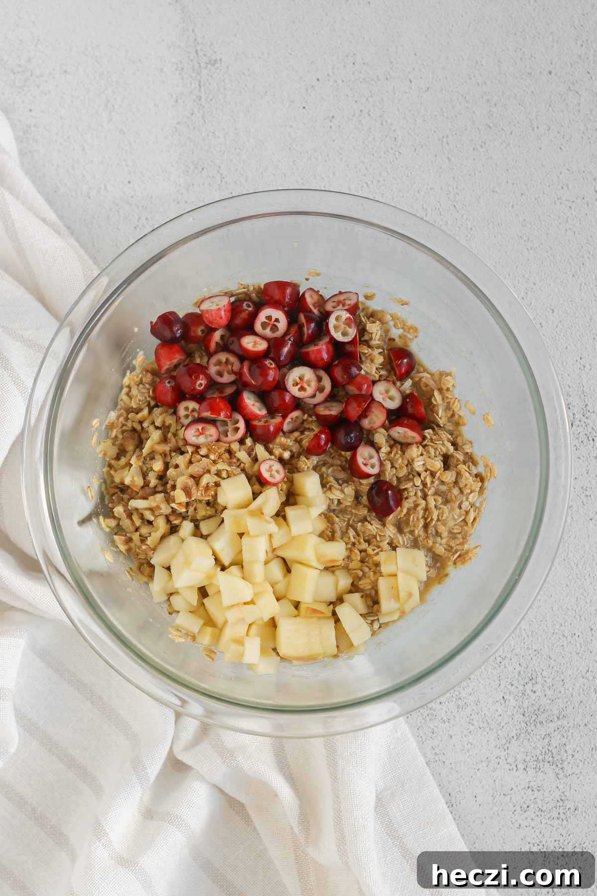 Oatmeal breakfast cookie dough in a glass mixing bowl with cranberries, apples, and walnuts added to the top.