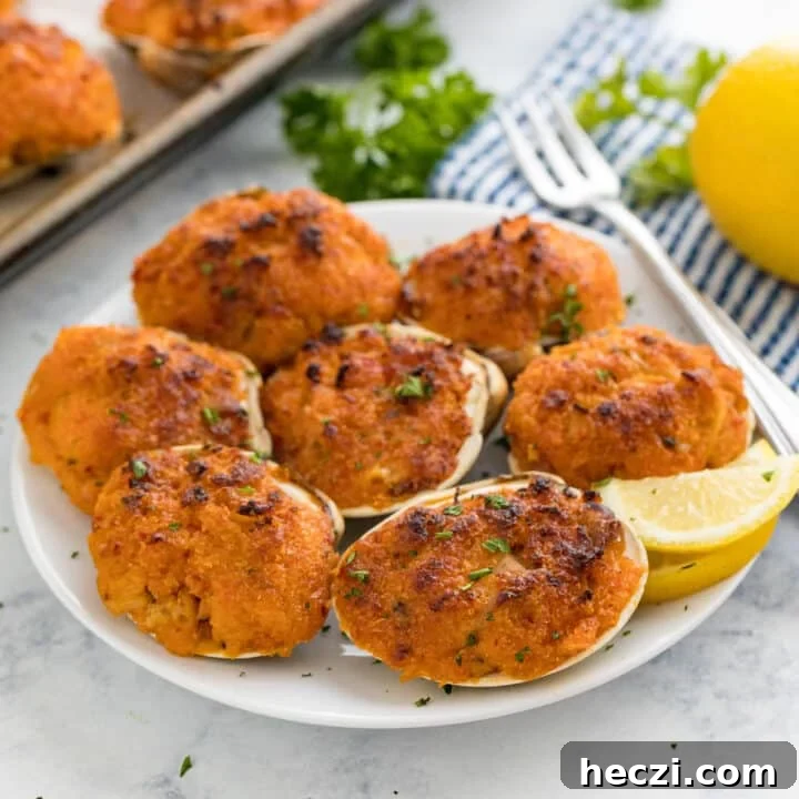 A plate of baked clams with a cocktail fork and lemon wedge on it with the pan, a striped napkin, lemon and parsley behind it.
