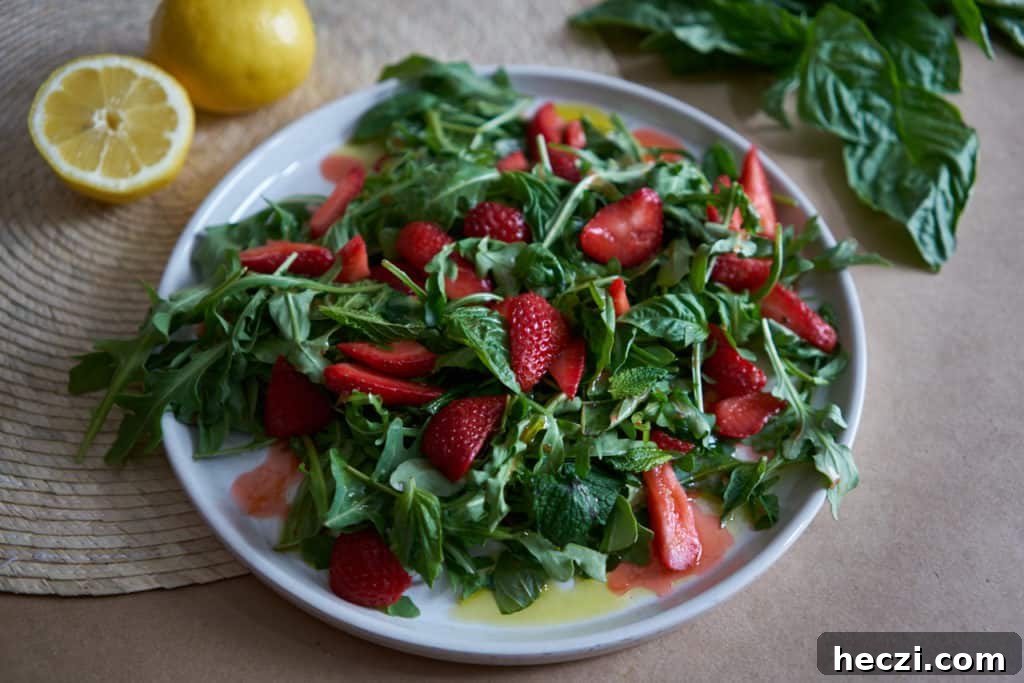 Spring Strawberry and Arugula Salad with Balsamic Glaze 2 A plate of salad with arugula, herbs, and sliced strawberries on a brown surface with basil leaves and lemons in the background.