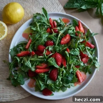 Spring Strawberry and Arugula Salad with Balsamic Glaze 6 A plate of salad with arugula, herbs, and sliced strawberries on a brown surface with basil leaves and lemons in the background.