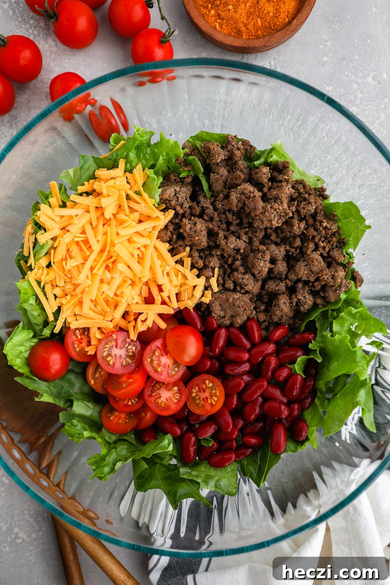 A large clear mixing bowl filled with fresh lettuce, ground beef, kidney beans, tomatoes and cheddar cheese sitting next to a small bowl of taco seasoning and cherry tomatoes on the vine