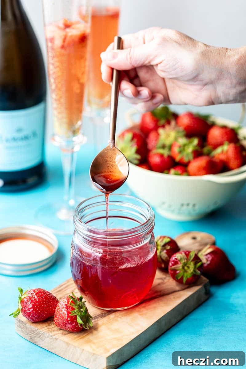 Homemade Strawberry Syrup Recipe for Drinks & Desserts 2 A hand holds a spoon with strawberry syrup over a jar; fresh strawberries, a bowl of strawberries, glasses of strawberry mimosas, and a prosecco bottle are in the background.