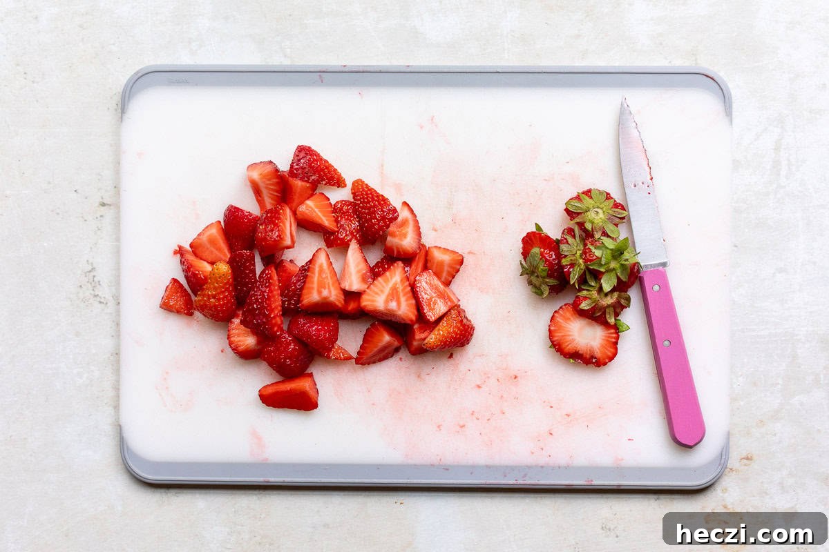 Homemade Strawberry Syrup Recipe for Drinks & Desserts 4 A white cutting board with chopped strawberries, strawberry tops, and a pink-handled knife.