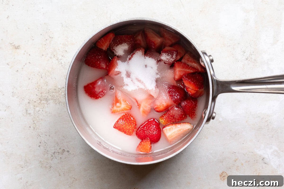 Homemade Strawberry Syrup Recipe for Drinks & Desserts 5 A saucepan containing chopped strawberries, sugar, and water on a light-colored surface.