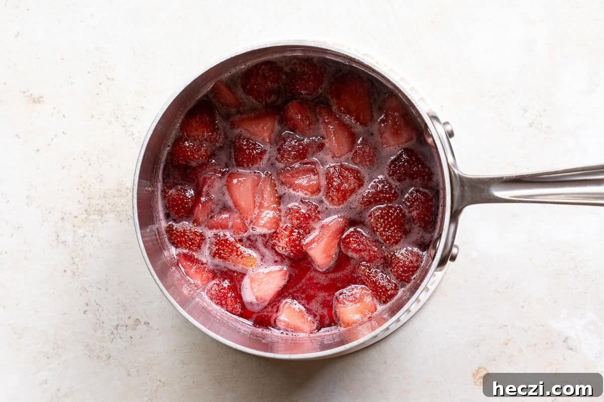 Homemade Strawberry Syrup Recipe for Drinks & Desserts 6 A stainless steel saucepan filled with strawberries simmering in a red liquid on a light-colored surface.