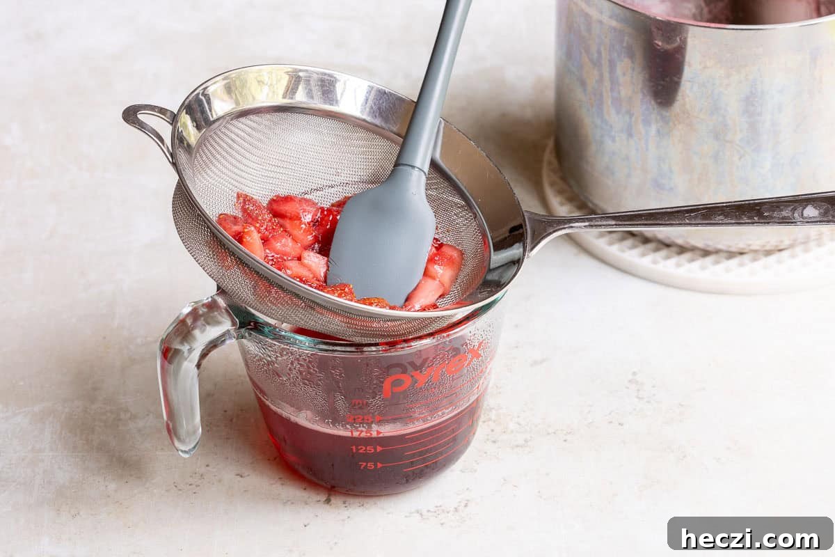 Homemade Strawberry Syrup Recipe for Drinks & Desserts 7 Sliced strawberries are being strained with a mesh sieve and spatula over a glass measuring cup, collecting strawberry syrup, with a pot in the background.