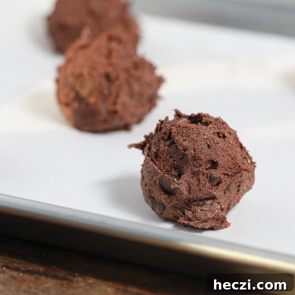 Ultimate Triple Chocolate Cookie Recipe Everyone Will Love 4 Scoops of rich triple chocolate cookie dough are placed on a parchment-lined baking sheet, ready to be baked. The dough is chunky and indulgent, filled with chocolate chips or chunks for the ultimate Triple Chocolate Cookies experience.