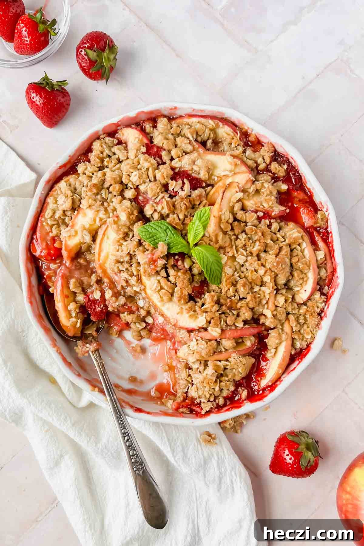 strawberry apple crisp baked to perfection with a golden crumble on top garnished with mint leaves and a spoon serving it.
