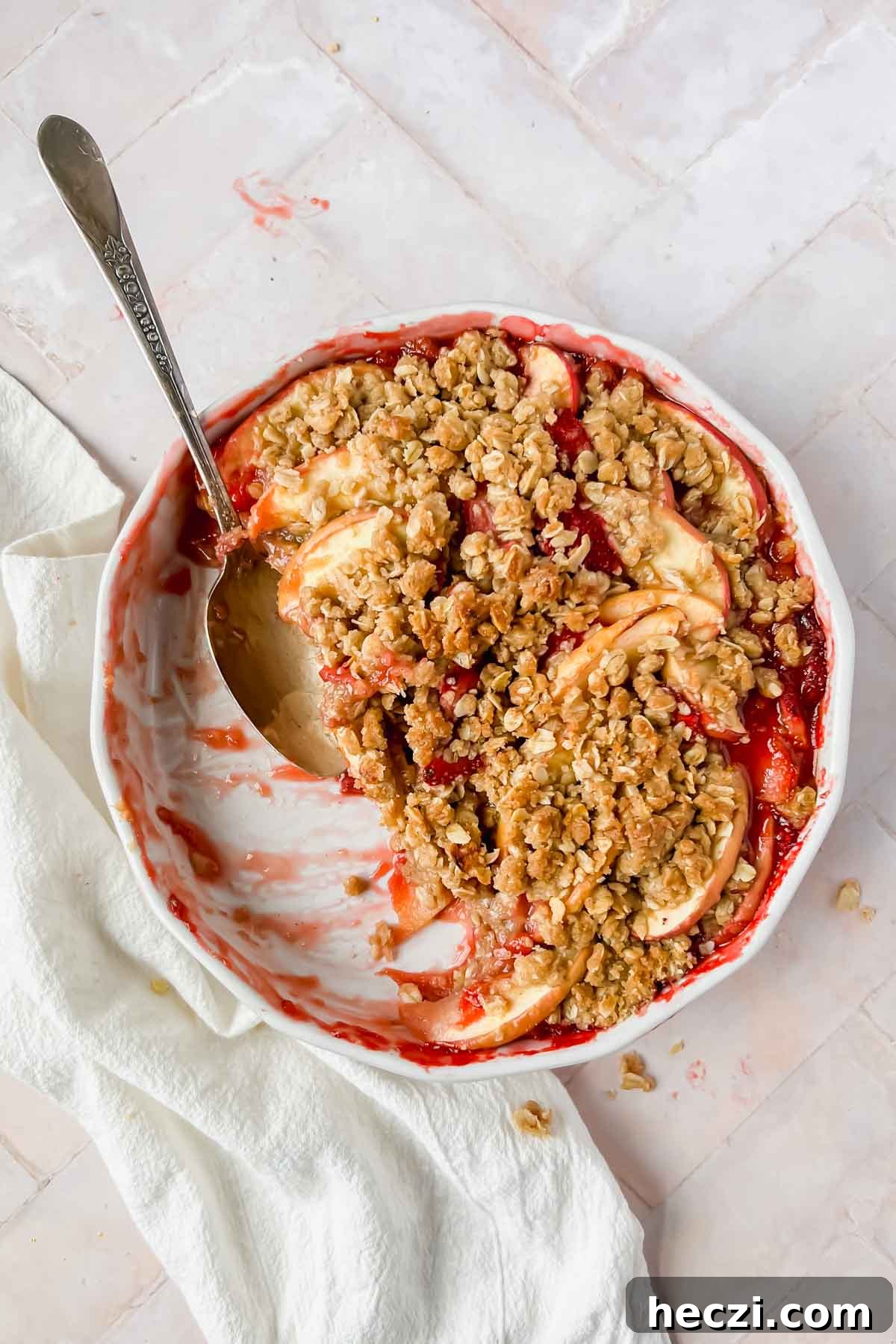 half eaten strawberry apple crisp with spoon on pink tiled background.