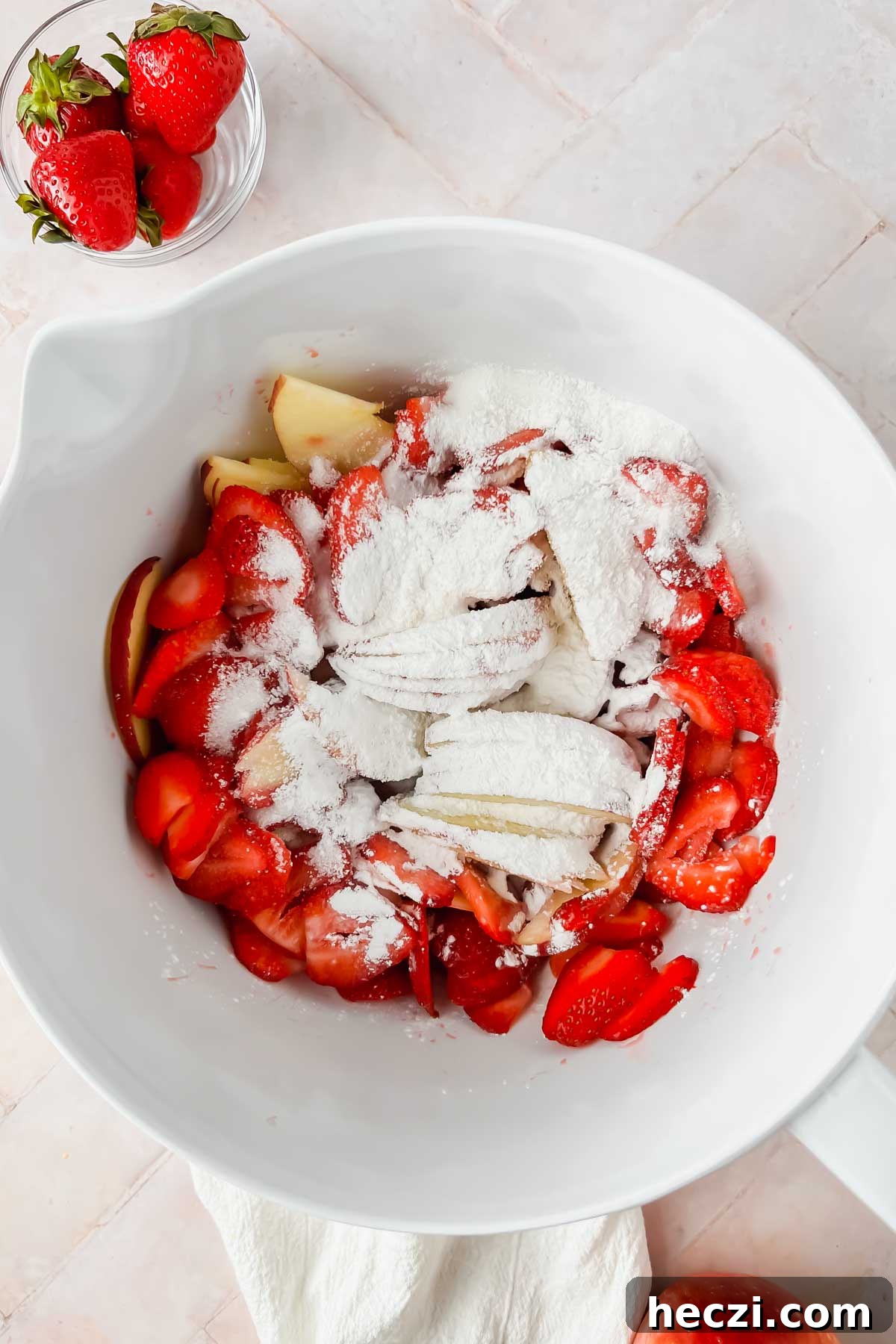 white mixing bowl filled with sliced strawberries, apples, tapioca flour, lemon extract and lemon zest.