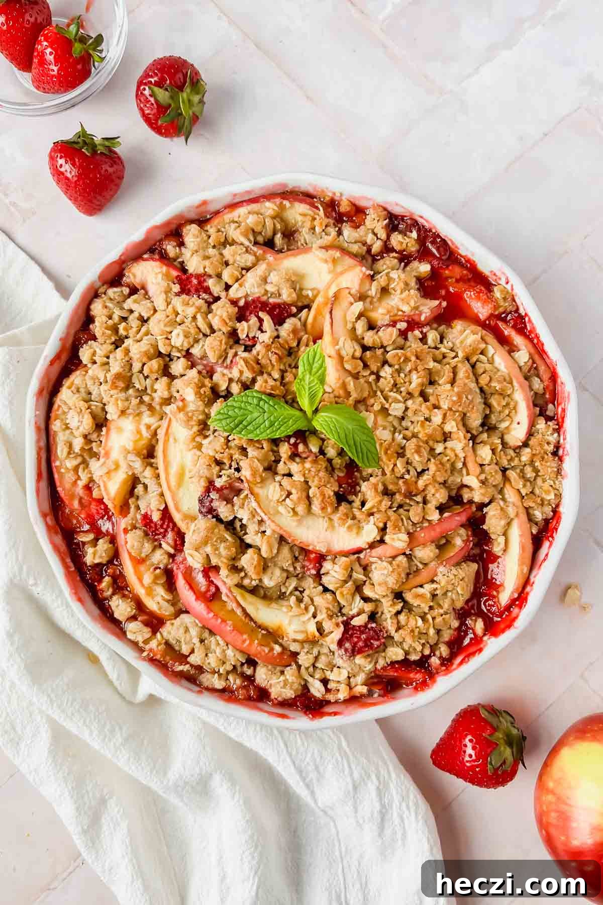 cooked strawberry apple crisp garnished with fresh mint and strawberries on pink tiled backdrop.