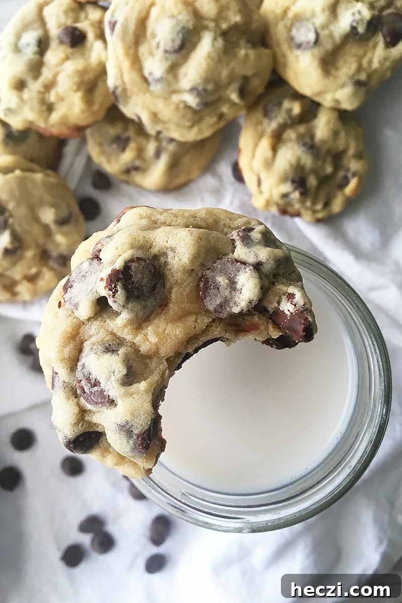 Vertical image of a half-eaten cookie on top of a glass of milk with more desserts in the background.