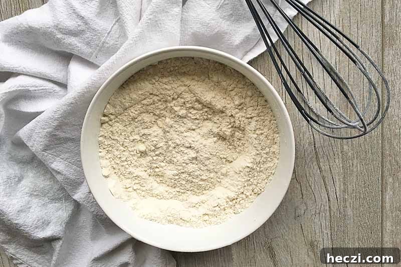 Horizontal image of a white dry mix in a white bowl next to a black whisk and white towel.