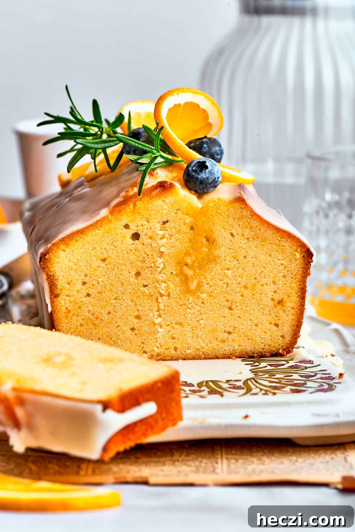 A side view of a slice of orange cake and the entire loaf, topped with blueberries, fresh rosemary, and orange slices.