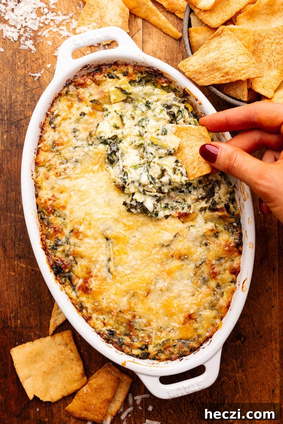 Creamy Baked Spinach and Artichoke Dip Recipe 5 A hand dipping a pita chip into a white oval dish with baked artichoke dip in it. Around the dip is a bowl of pita chips.