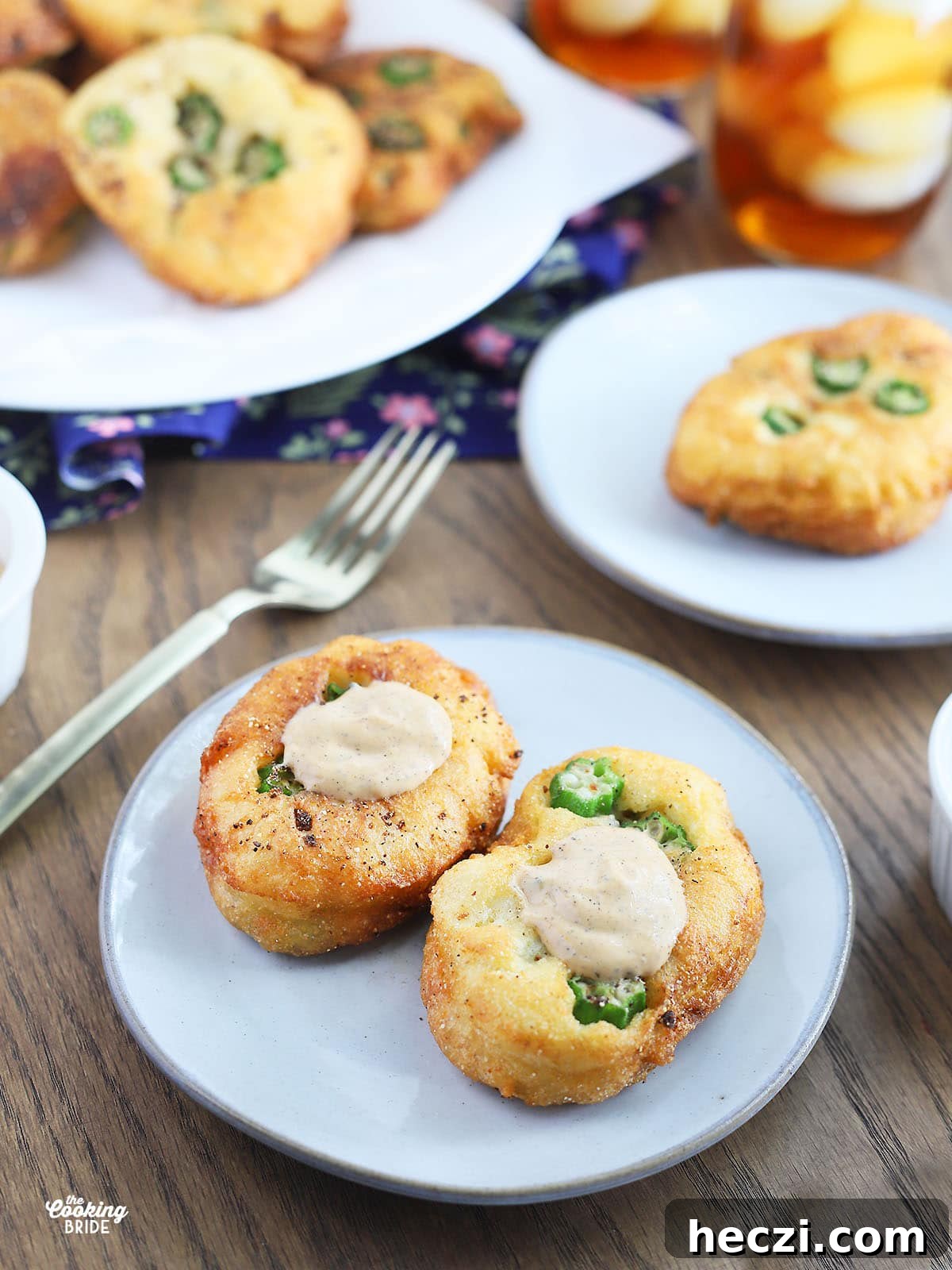 side shot of two okra hoe cakes with sauce on a white plate. A gold fork and a platter of hoe cakes are in the background.