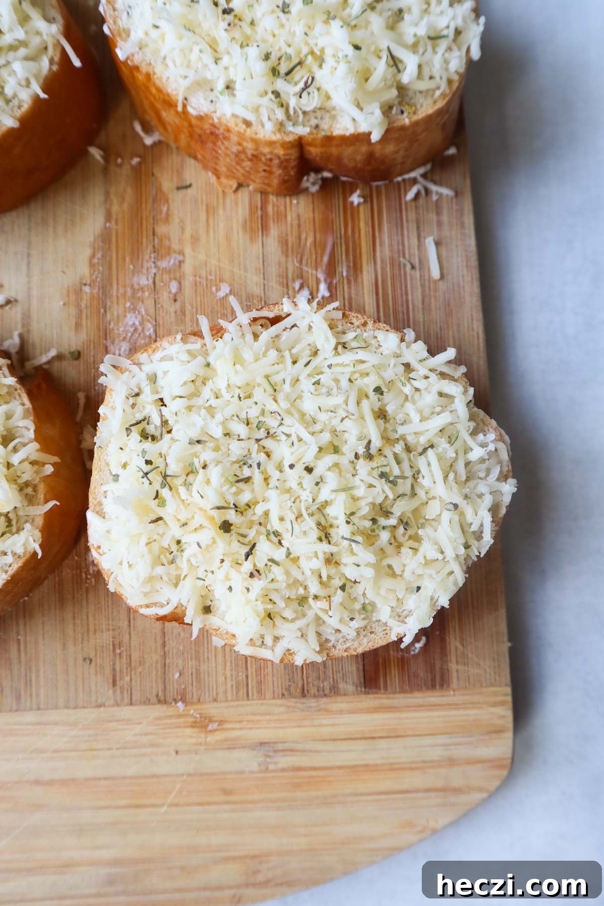 wooden cutting board with one slice of french bread topped with garlic herb butter and shredded cheese with a dash of italian seasoning