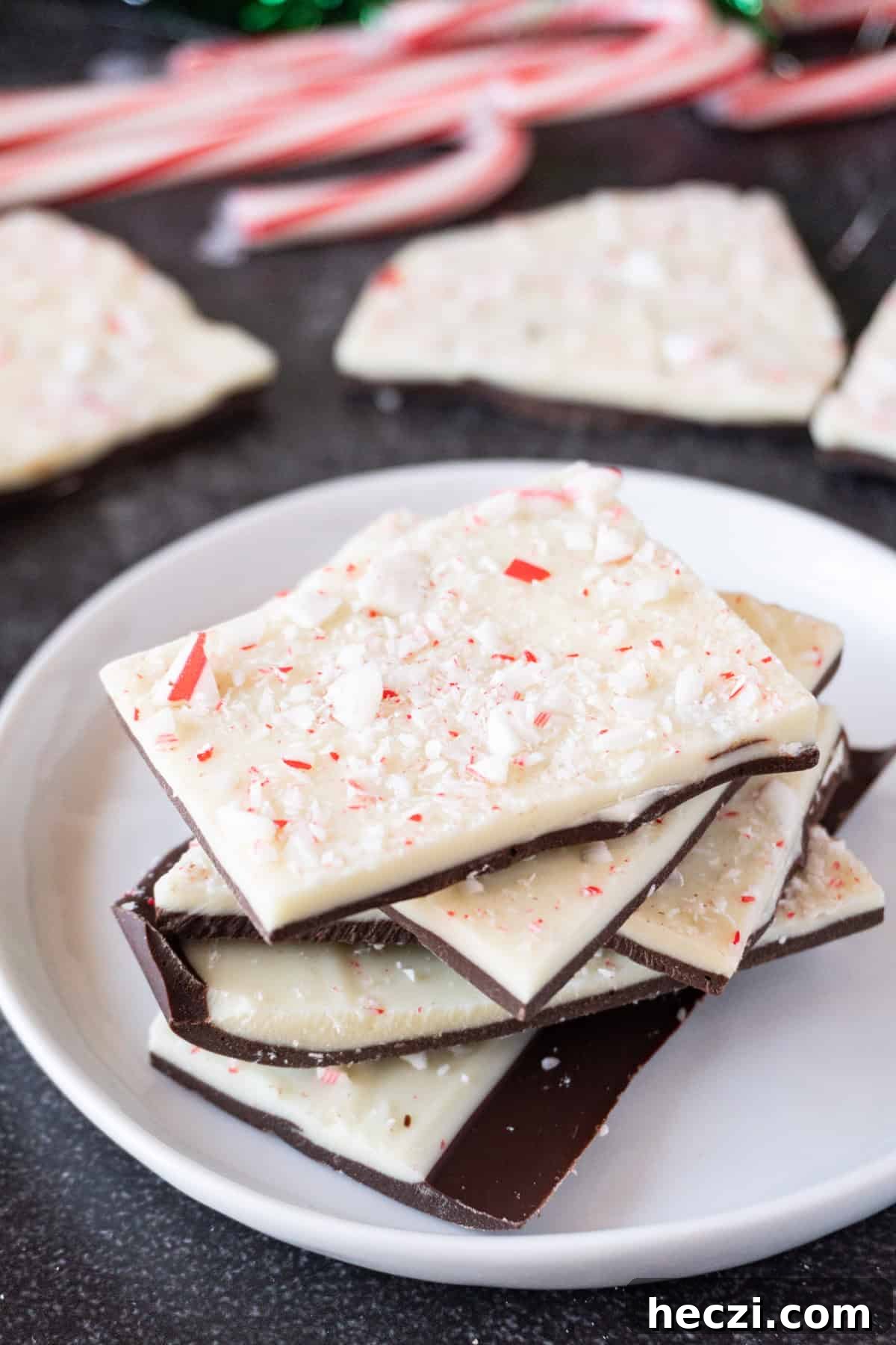 Homemade Peppermint Bark Recipe: Quick Holiday Chocolate Candy 2 Peppermint bark stacked on a white plate with more peppermint bark and candy canes in the background.