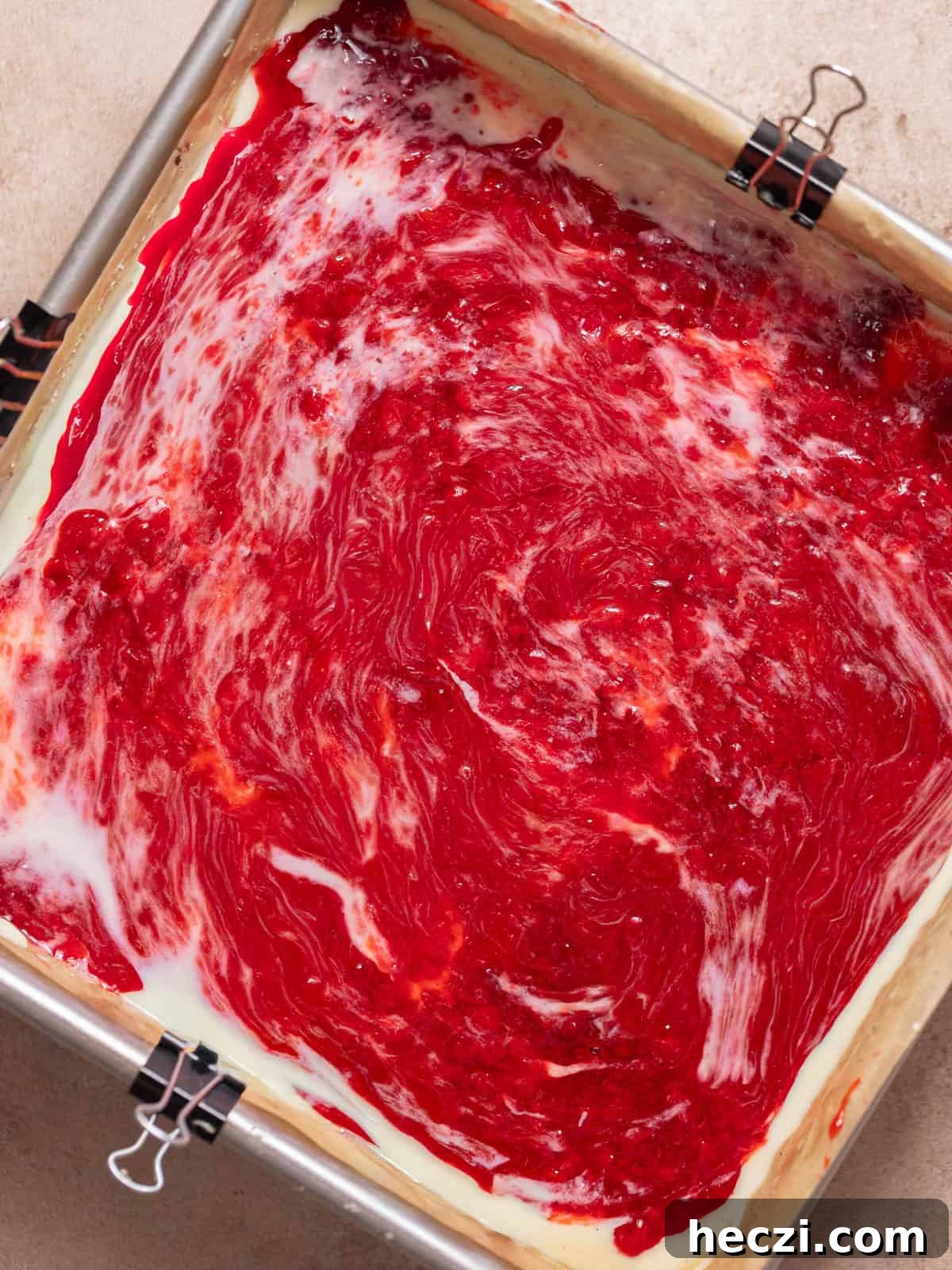 Strawberry jam swirled over cheesecake filling in a square pan.