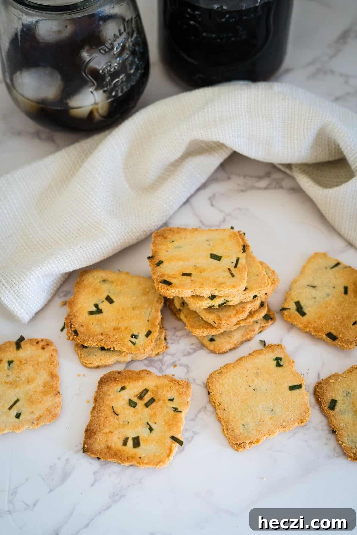 Cottage Cheese Crackers Recipe: Crispy Low-Carb Snack Guide 2 A stack of square, herb-topped crackers on a marble surface beside a white cloth and two jars filled with a dark beverage.