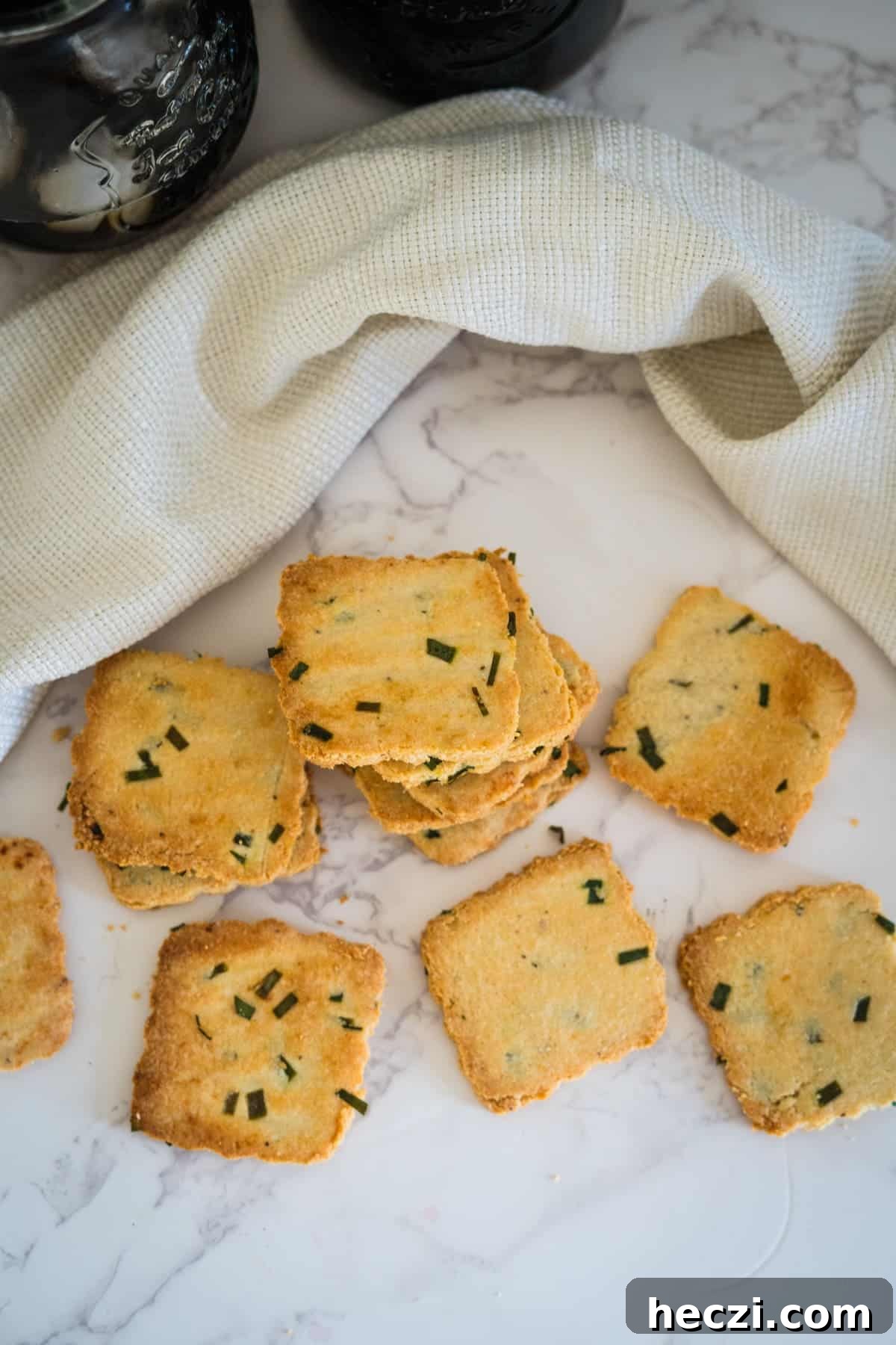 Cottage Cheese Crackers Recipe: Crispy Low-Carb Snack Guide 11 A stack of square, golden-brown cottage cheese crackers with green herbs sits on a marble surface next to a cloth and two jars.