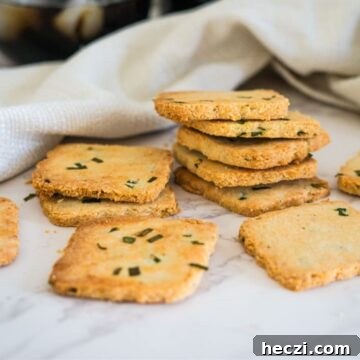 A stack of square cottage cheese crackers with green herbs rests on a white surface next to a white cloth. Some crackers are spread around in the foreground.