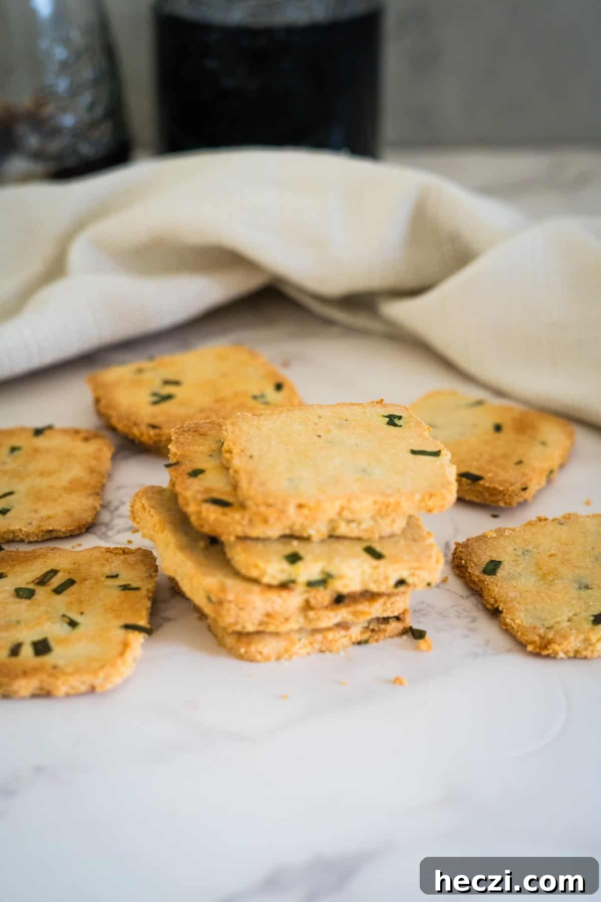 Cottage Cheese Crackers Recipe: Crispy Low-Carb Snack Guide 3 A stack of square, golden-brown cottage cheese crackers with green herbs are placed on a marble surface, with a white cloth in the background.