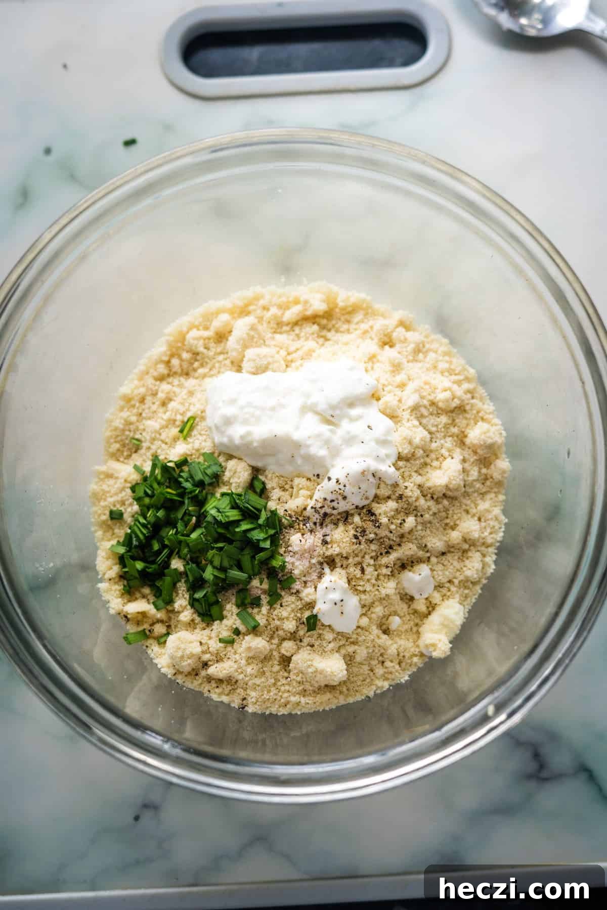 Cottage Cheese Crackers Recipe: Crispy Low-Carb Snack Guide 5 A glass bowl contains a mixture of almond flour, chopped green herbs, black pepper, and dollops of white cream or yogurt, placed on a marble countertop near a silver spoon.