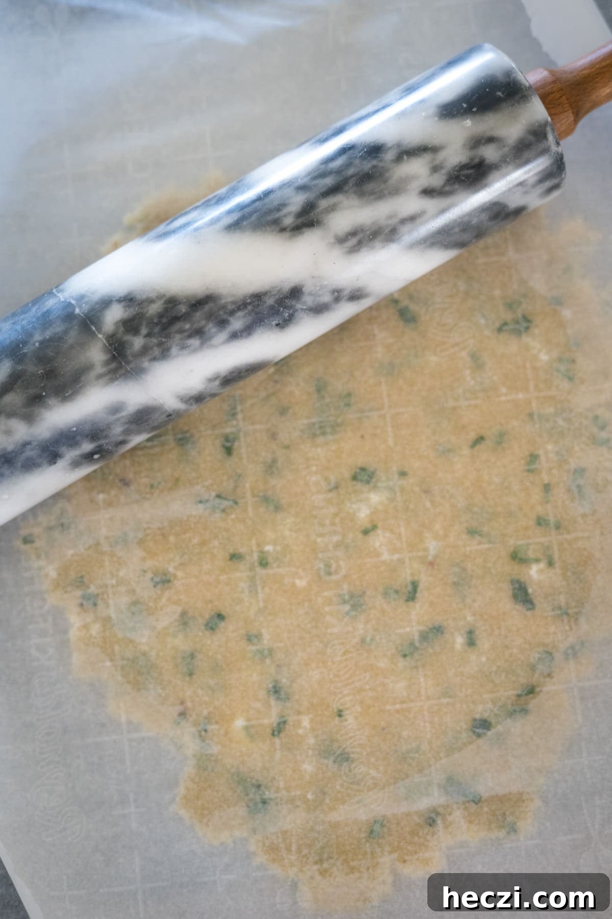Cottage Cheese Crackers Recipe: Crispy Low-Carb Snack Guide 7 A marble rolling pin rolls out a sheet of dough with green herbs, placed on parchment paper.