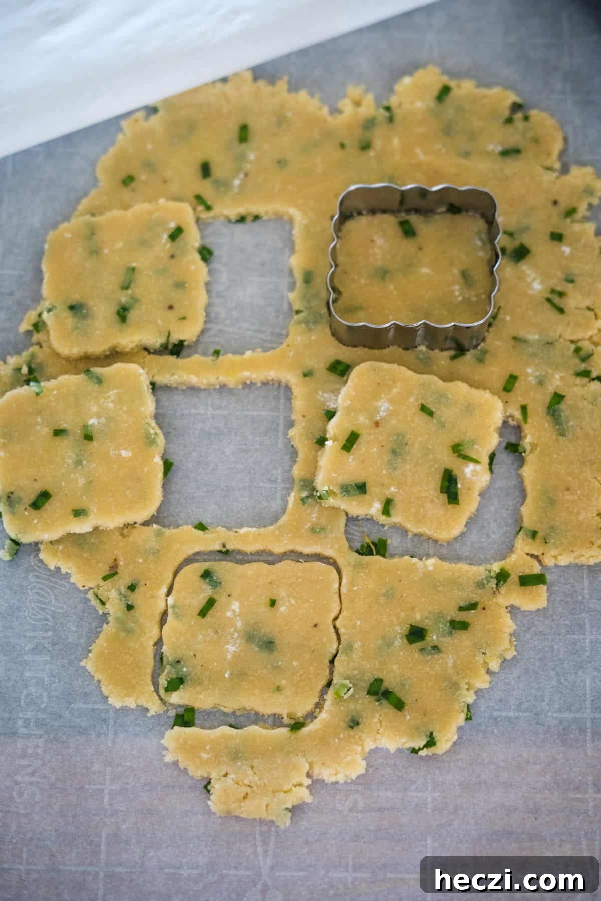 Cottage Cheese Crackers Recipe: Crispy Low-Carb Snack Guide 8 A sheet of rolled-out dough with green herbs, partially cut into square shapes using a square cookie cutter, laid on parchment paper. Some cut-out squares are arranged around the dough.