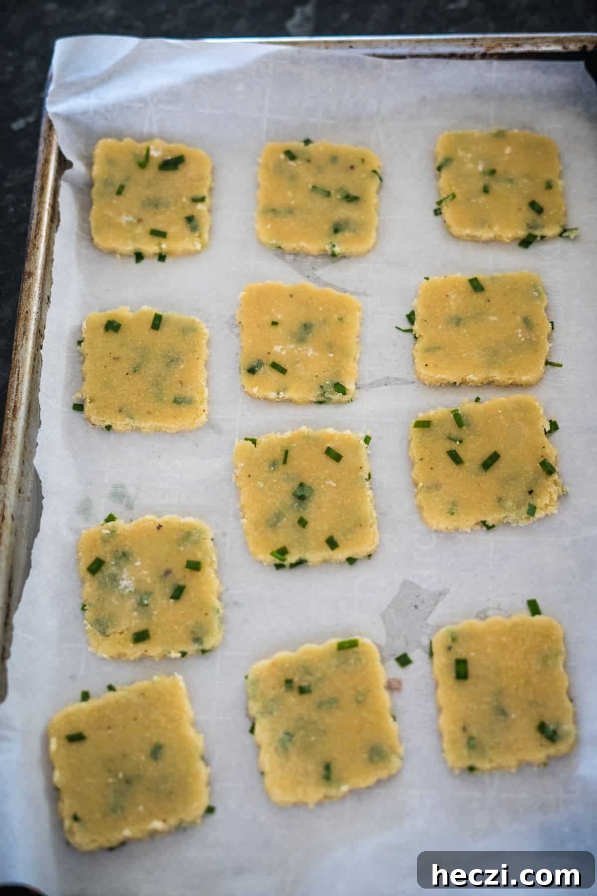 Cottage Cheese Crackers Recipe: Crispy Low-Carb Snack Guide 9 A baking tray with 12 uncooked square crackers dotted with green herbs, arranged in three rows, placed on parchment paper.