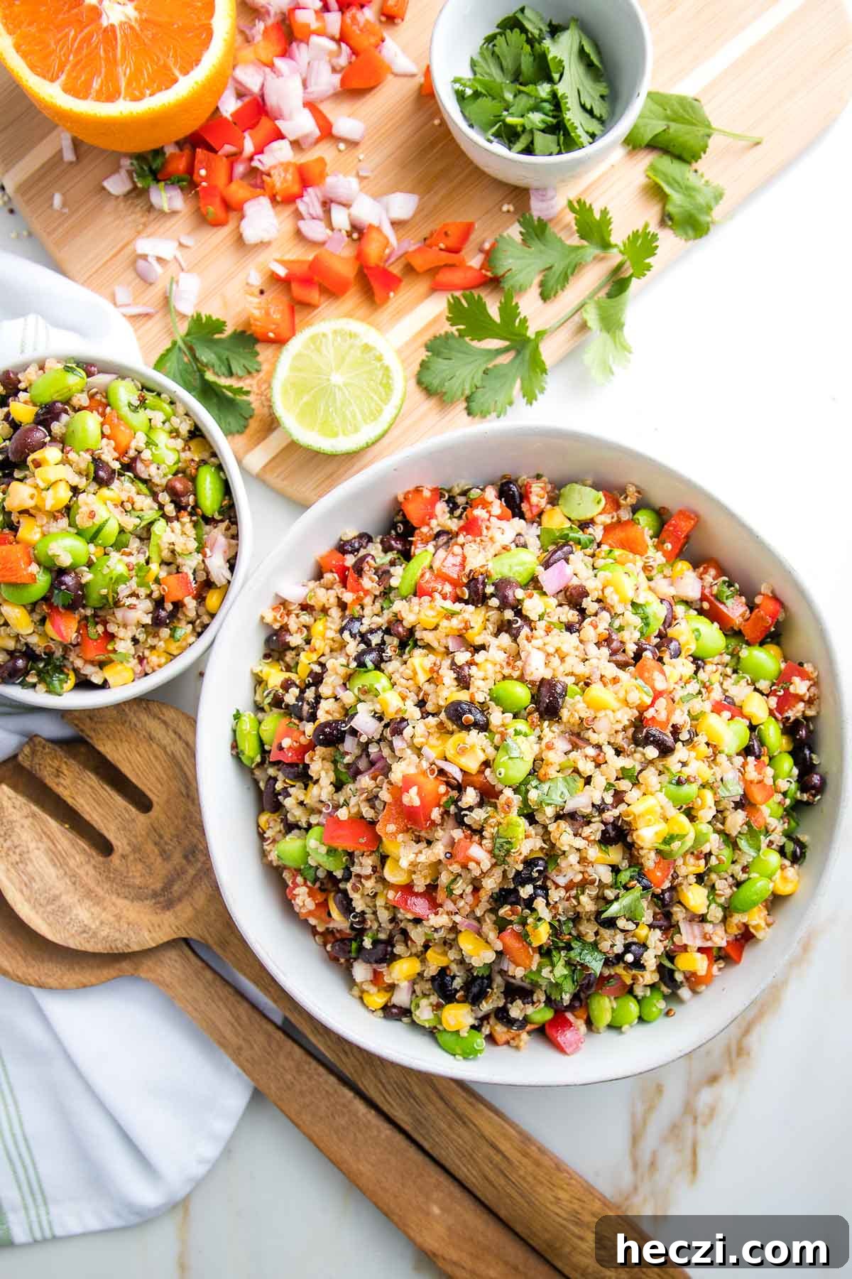 Large and small white bowls filled with colorful quinoa salad. In the background there is a cutting board with scraps of chopped red pepper, shallot and fresh cilantro. Also there is half an orange and half a lime. Wooden salad servers are next to the bowls.