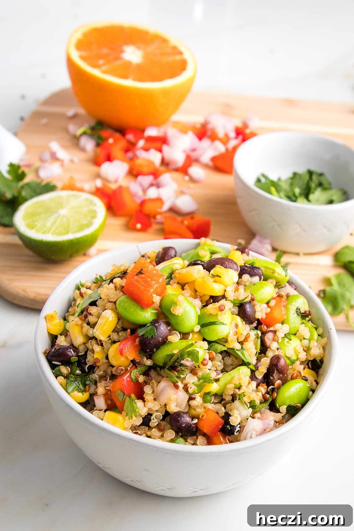 Bowl of colorful veggies plus quinoa and in the background a cutting board with half an orange, half a lime, chopped red peppers and shallots and cilantro
