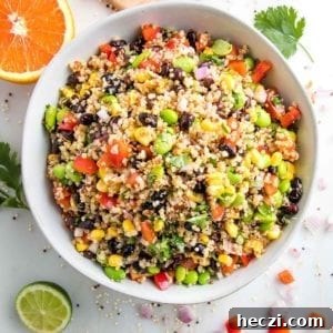 close up photo of a large white bowl filled with colorful quinoa salad that includes red peppers, green edamame, black beans, bright yellow corn and chopped cilantro. There is an orange half and a half a lime, plus fresh cilantro leaves around the bowl.